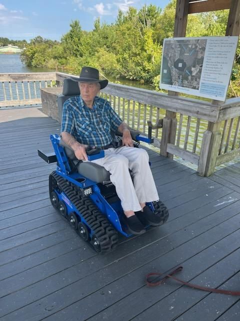 Man in khaki pants and plaid shirt sitting in tracked chair on dock at Trout Lake. 