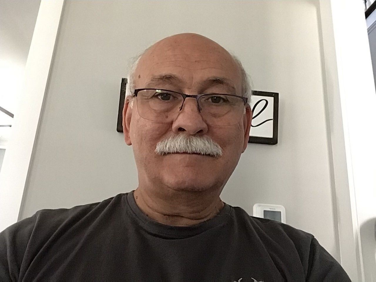A selfie-style image of Gary Caruso, an older gentleman with a thick moustache and glasses who is wearing a black shirt.