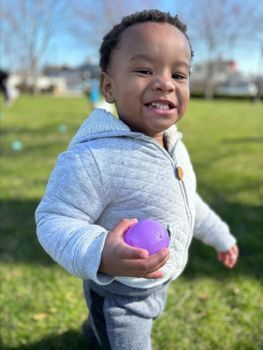 A toddler hanging out outside, holding a purple rubber ball and smiling.