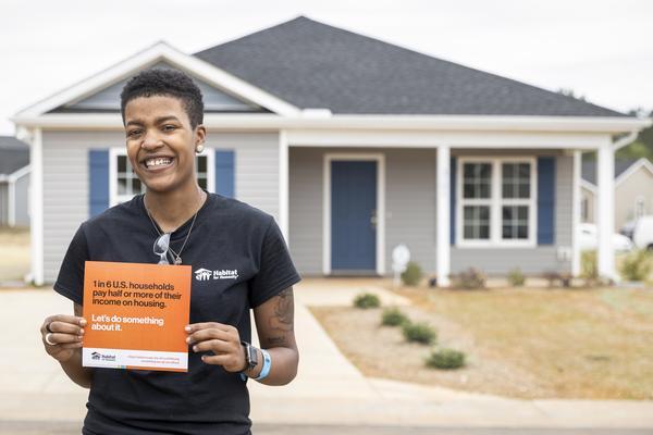 A woman stands in front of a new home, holding a Habitat for Humanity flyer that calls attention to the issue of housing affordability in the U.S. Herr bright smile reflects pride and hope.