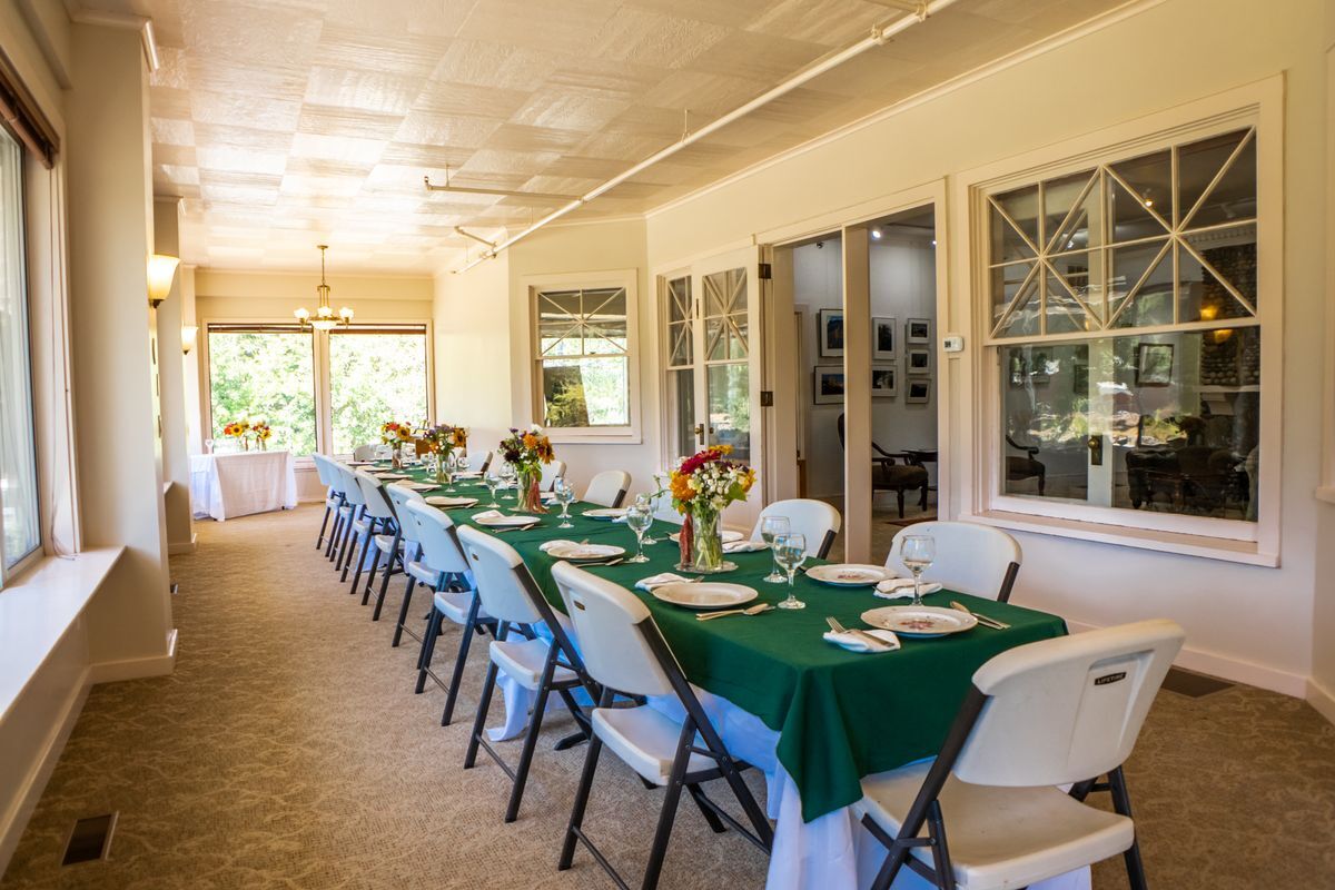 An image of the Sun Room set up dinner style with long tables, green table cloths, and dinner settings, surrounded by white chairs.