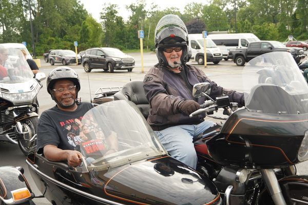 A man sits in a motorcycle sidecar wearing a helmet and smiling at a camera while a rider sits in the motorcycle next to him, also facing the camera