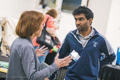 A man with dark, curly hair stands talking to a woman. You see her back. Her arms are gesturing as she talks.