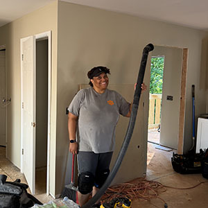Volunteer in Clemson Veterinary Medicine shirt smiling while holding a shop vacuum hose in a home under renovation
