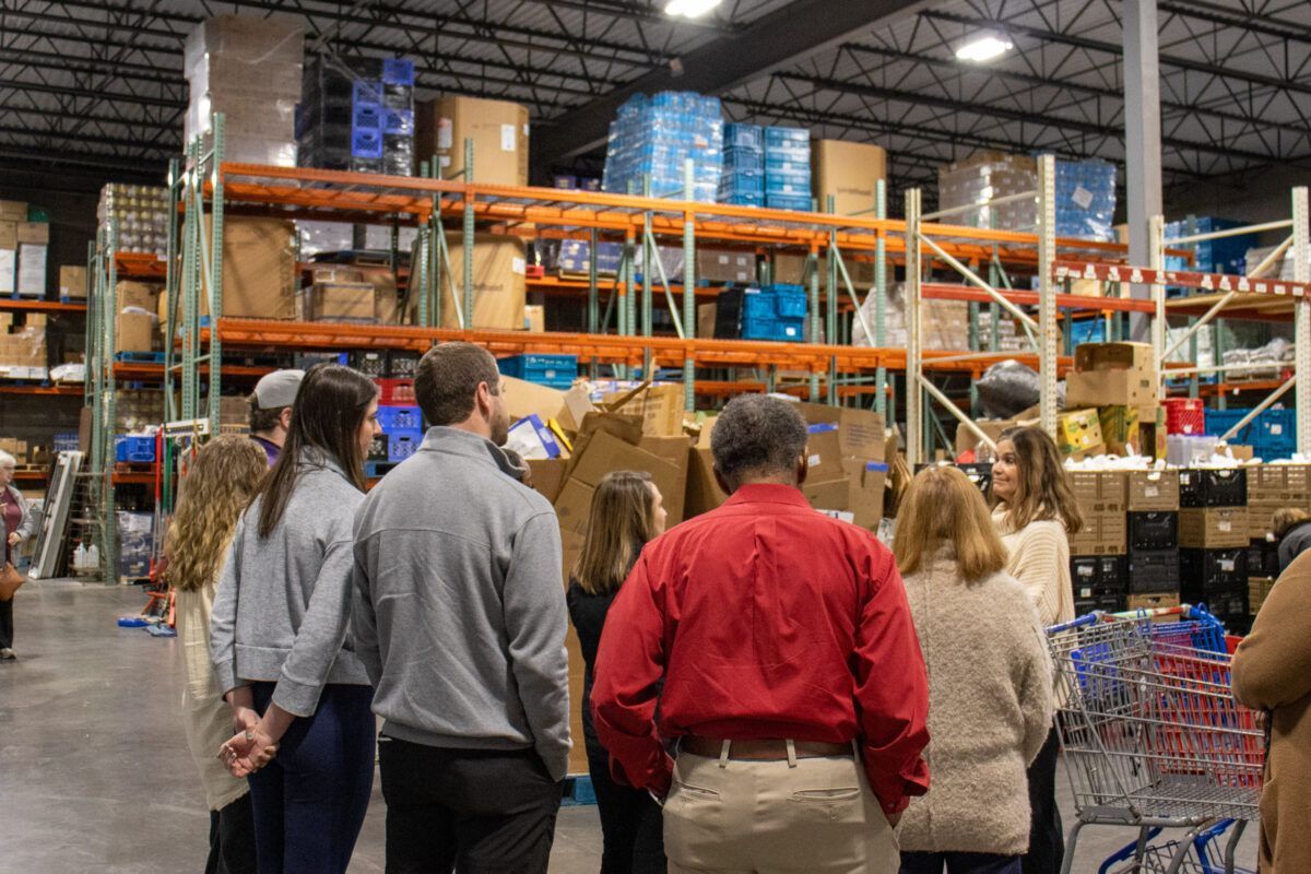 A group of people in a warehouse listening to a woman speaking, surrounded by shelves stacked with boxes and crates.