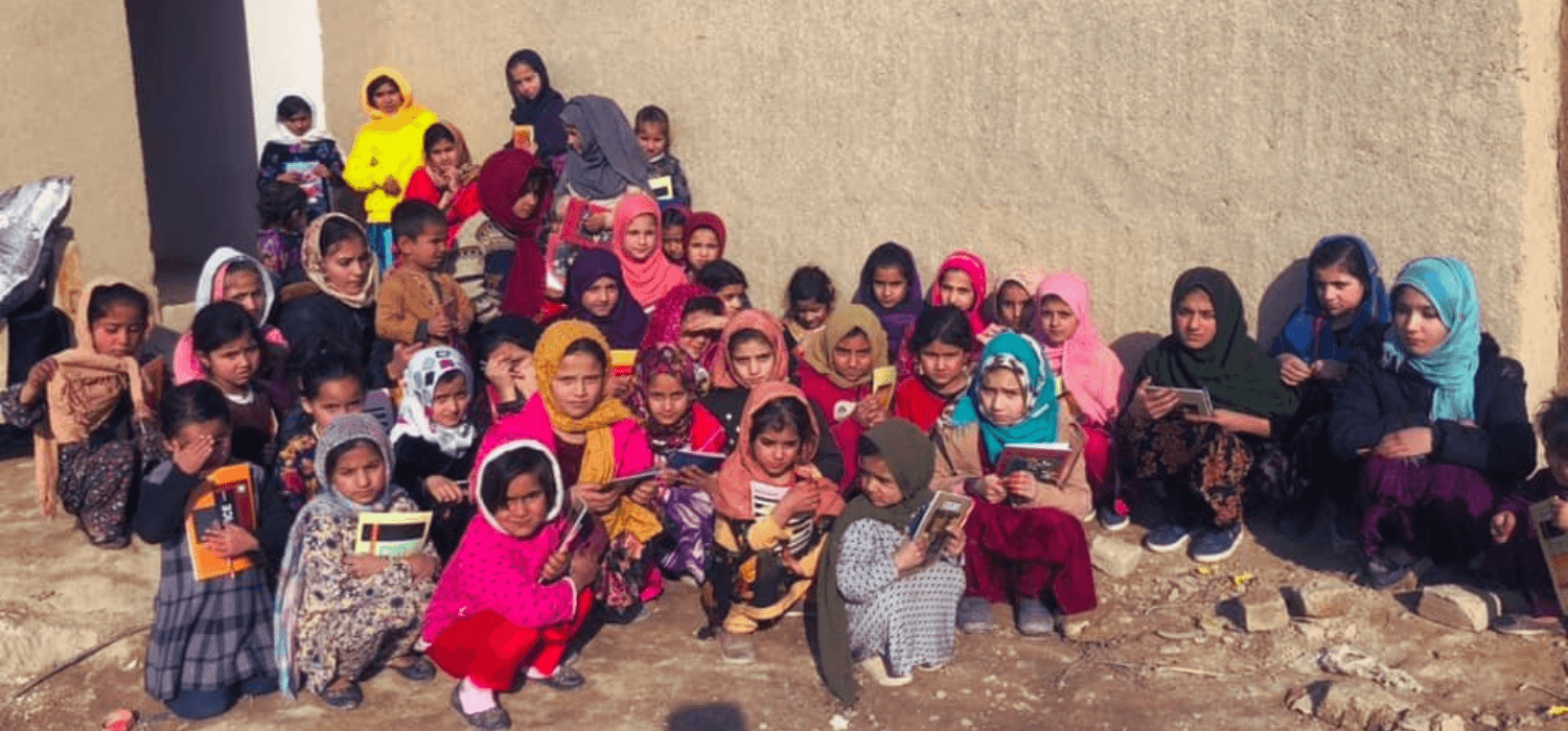 A large group of Afghan girls squatting on the ground and crouching against a wall, holding textbooks. 