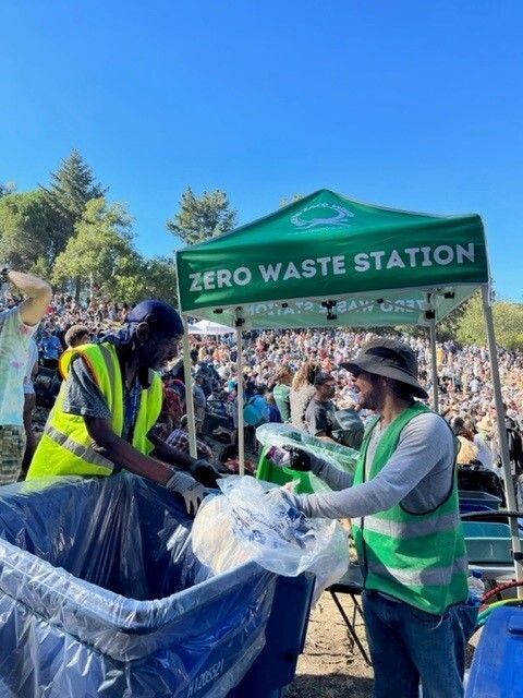 A black man and a white man in safety vests talk and sort waste at bins in front of a Zero Waste Station in front of a large crowd at a festival type event