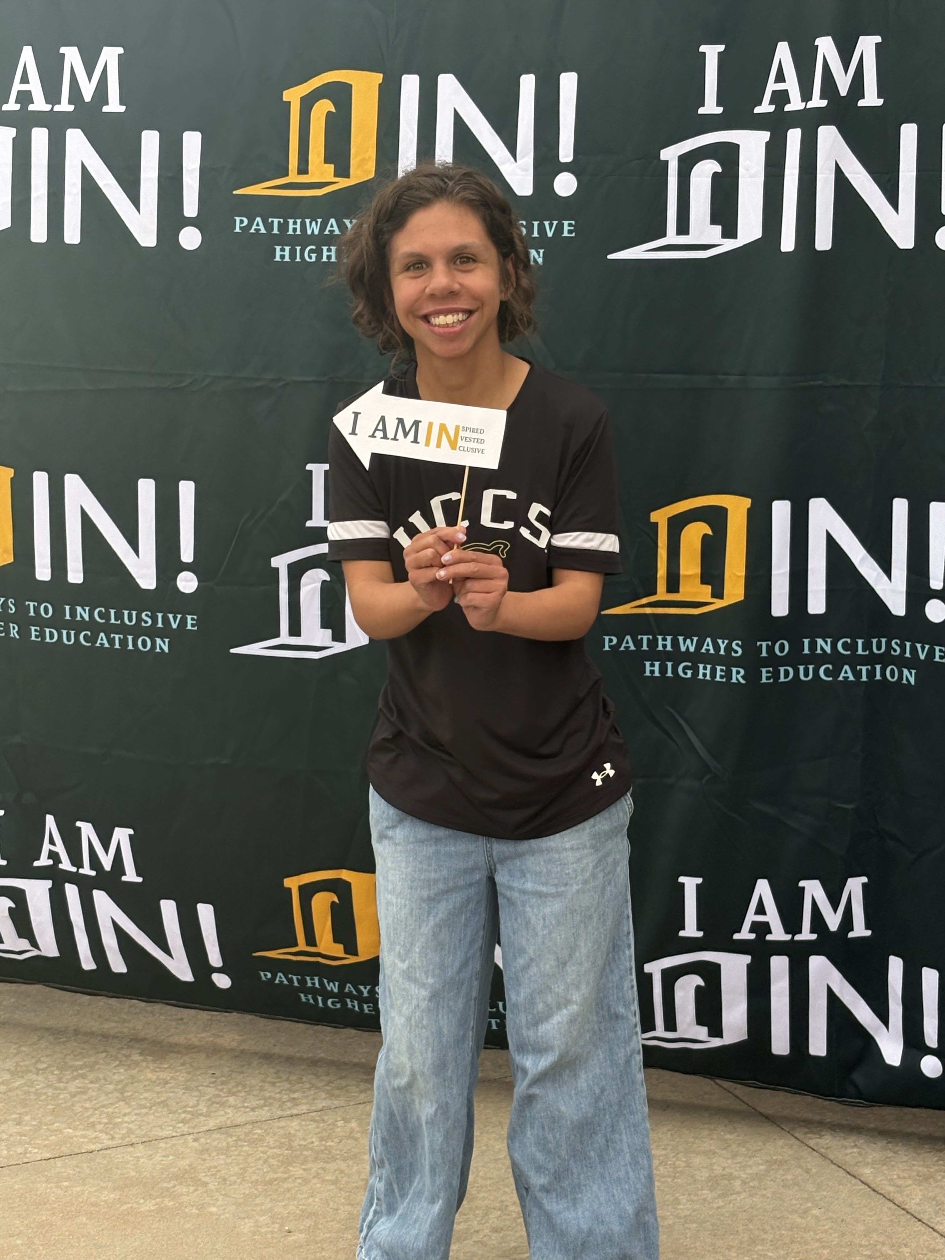 Maggie wears a black UCCS t-shirt and jeans. She holds a white arrow which says, "Be INspired, INvested, INclusive."