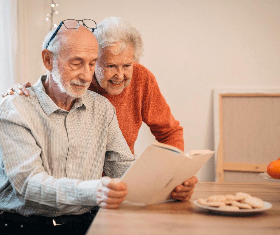 An elderly couple sits at a kitchen table reviewing a document together. The man holds the paper while the woman leans over his shoulder smiling, with a plate of cookies and soft lights in the background creating a warm, homey setting.