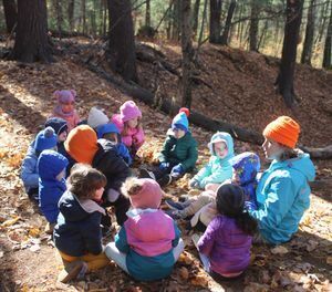 A circle of preschool children sitting in the woods listening to nature and science educator. 