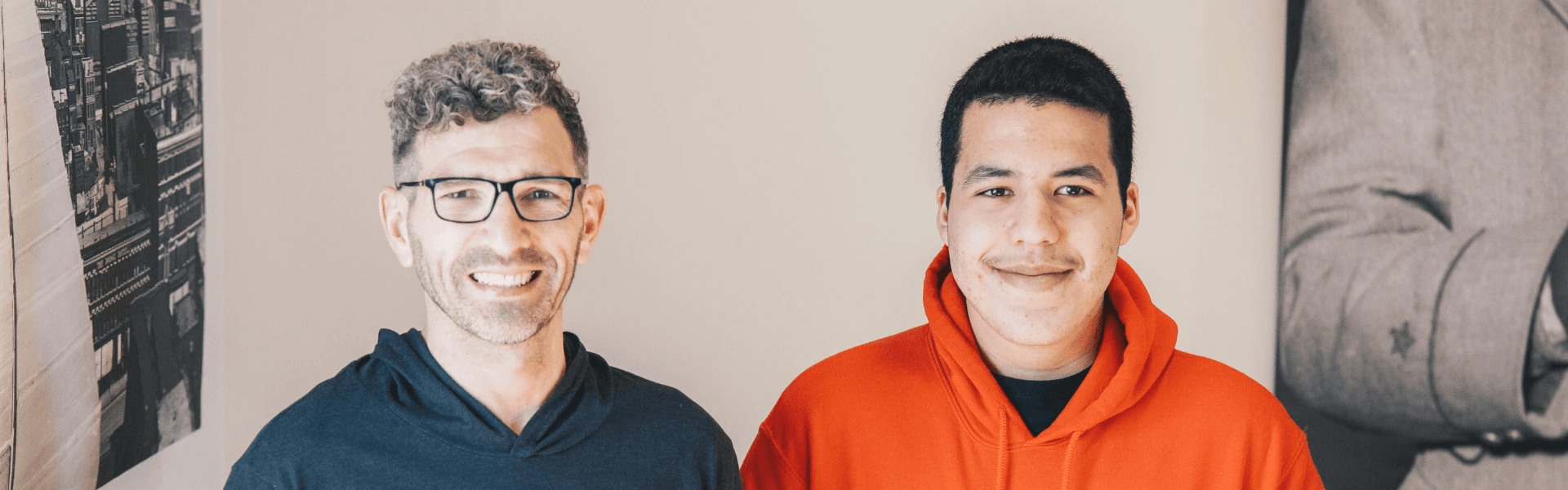 A mentor and mentee from Big Brothers Big Sisters smile together indoors; the mentee, wearing a bright red hoodie, stands in the foreground while the mentor, wearing glasses and a dark shirt, stands slightly behind him.