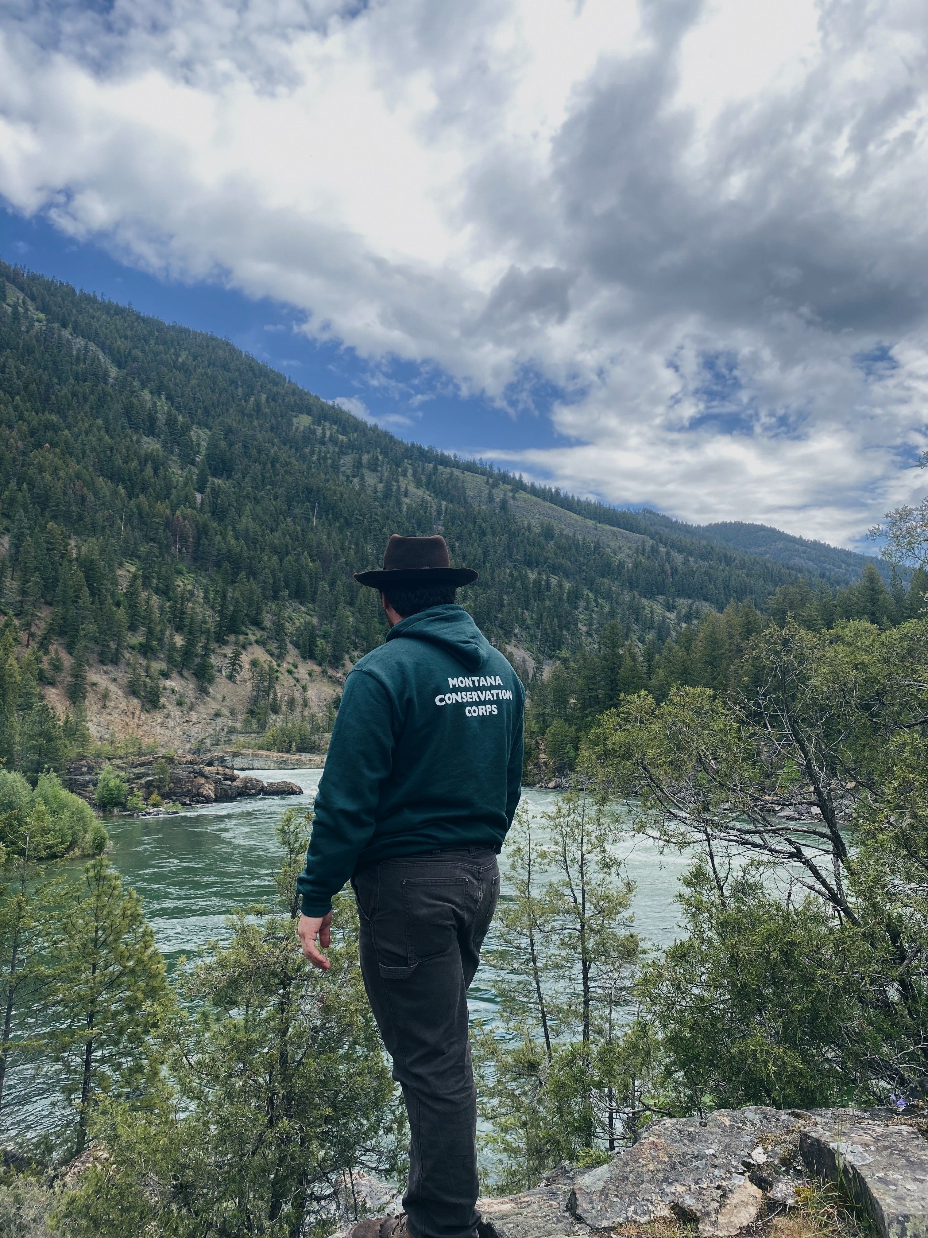 A person wearing a black cowboy hat stands with their back to the camera, overlooking a beautiful river and forested mountainside.