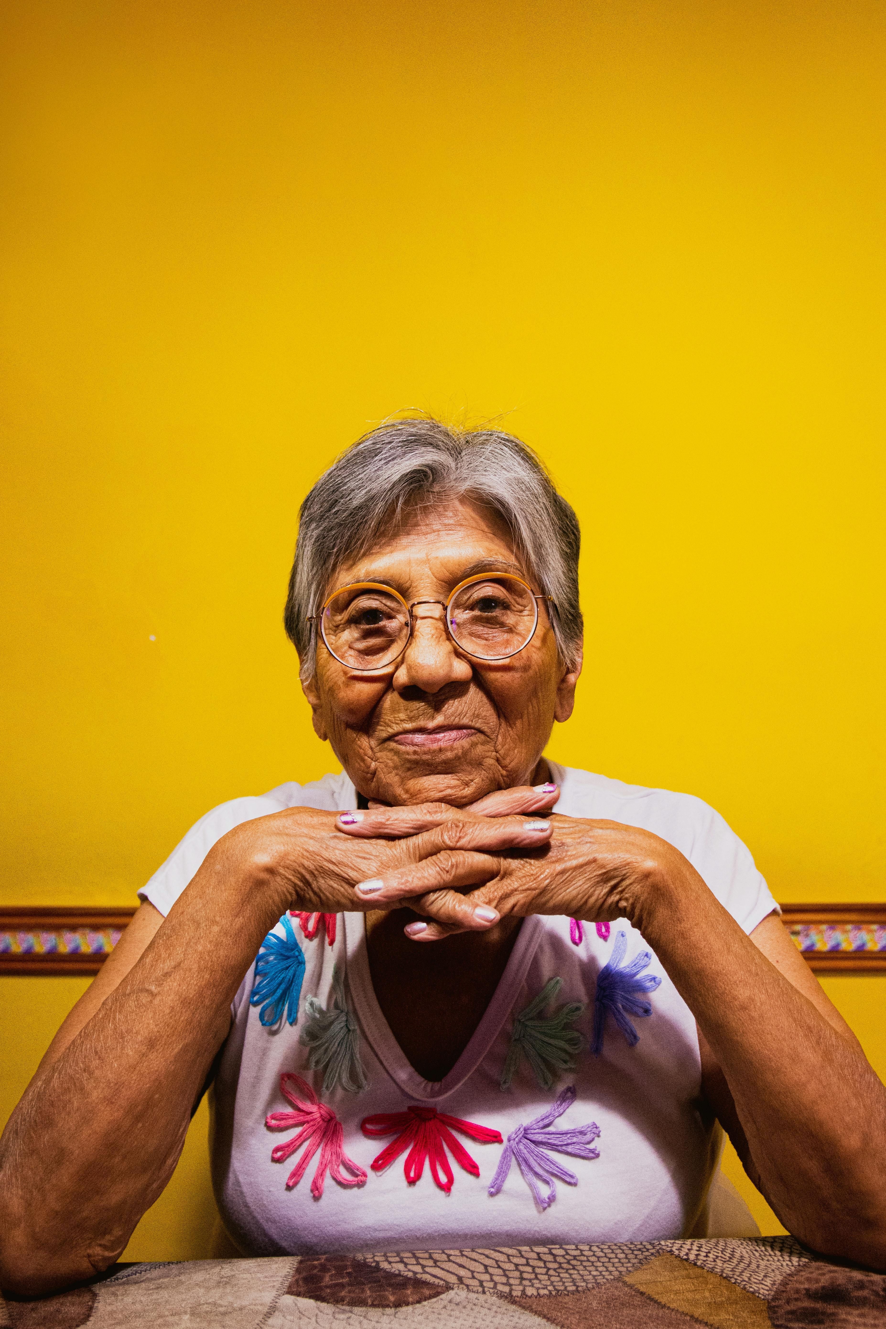 A woman is sitting at the table with her hands linked underneath her chin. She is wearing a light purple shirt with decorative flowers on it. The wall behind her is bright yellow. 