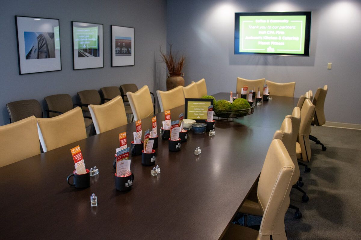 A conference room with a long table, beige chairs, and decorative items. A screen on the wall displays a message.