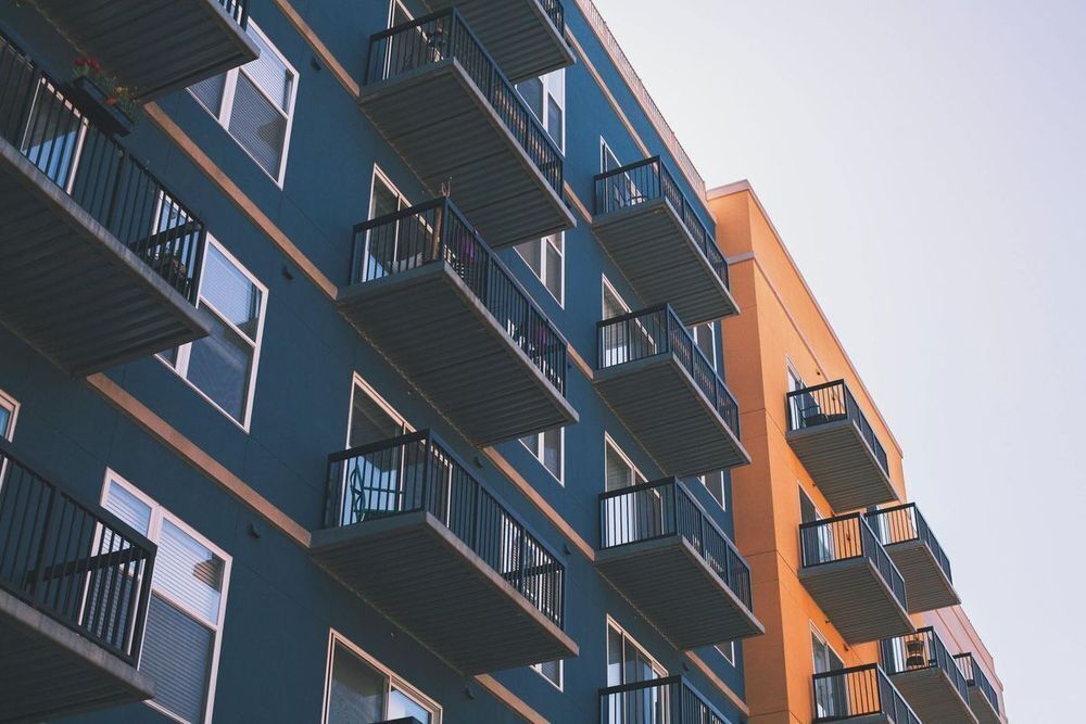 A dark blue apartment building is next to an orange apartment building. Windows and balconies are visible with a small section of gray sky at the top right of the image.