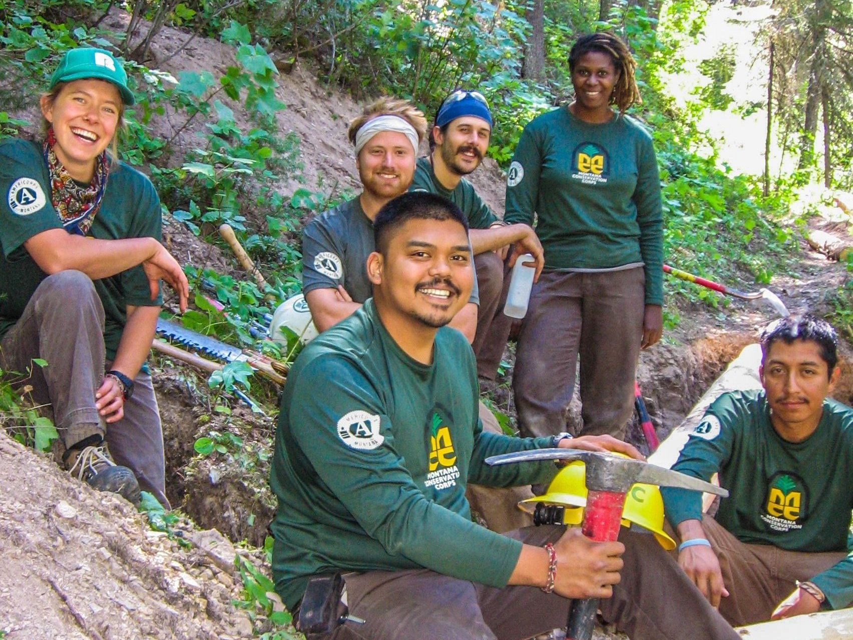 [Image description: Six MCC members, all sitting along a hillside smiling. Tools, helmets, hand saws and foliage surround them. They look as if they are in the midst of a lunch break, finishing up a drainage feature on a trail they are maintaining.]