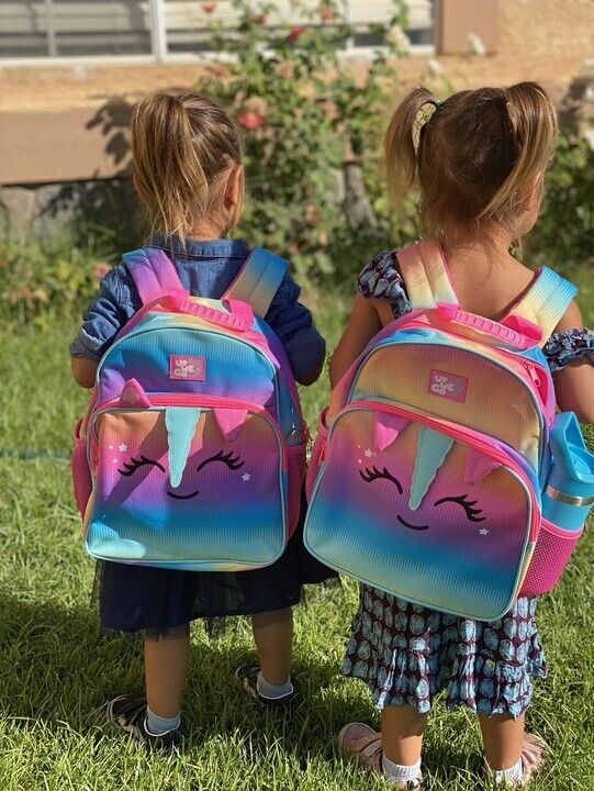 Two small children standing on grass, carrying rainbow-colored backpacks.
