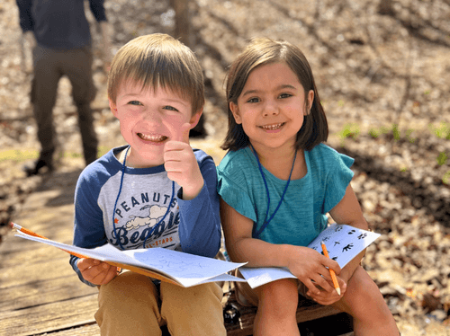Two sitting smiling kids holding their nature journals