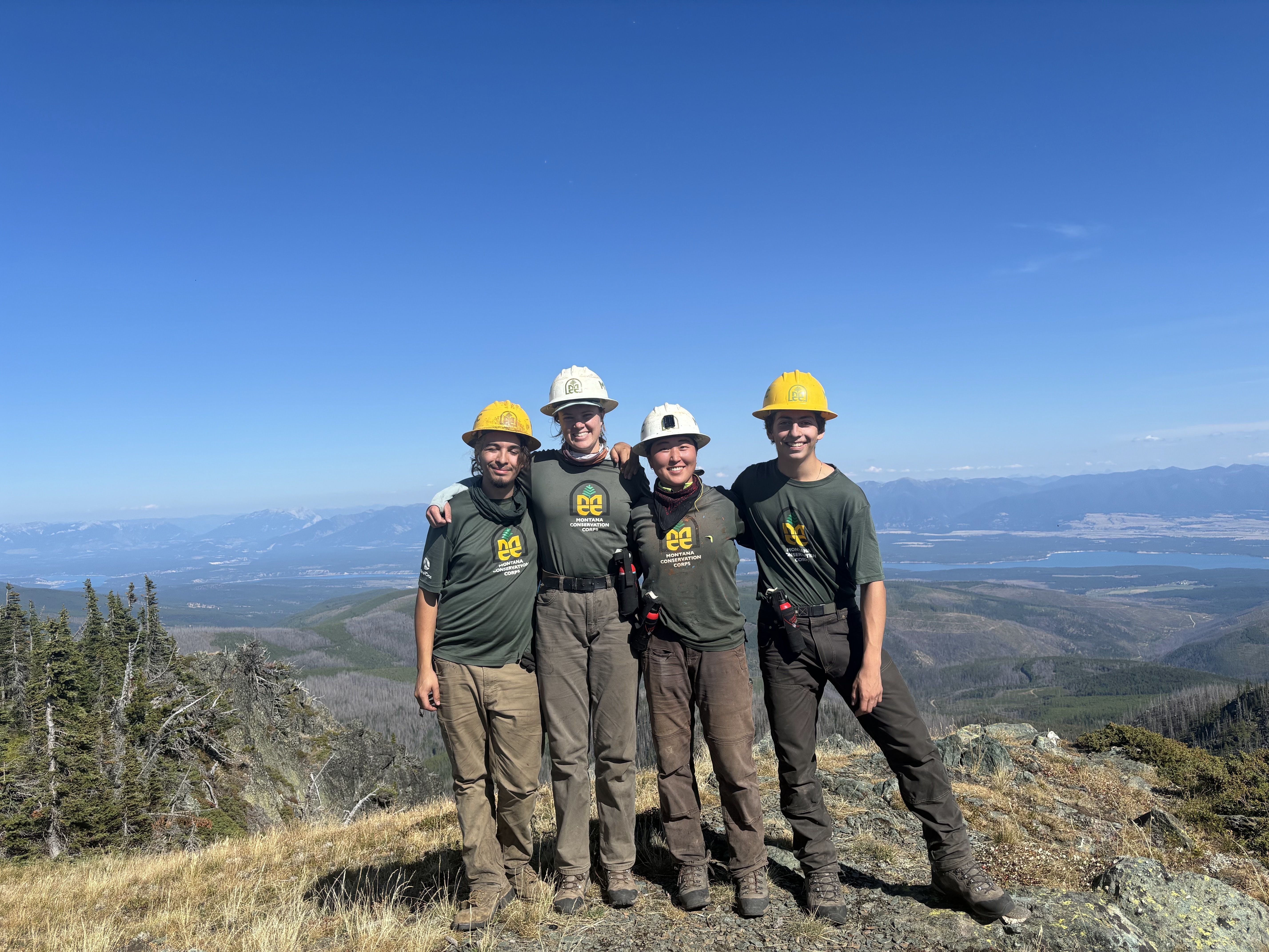 A crew stands on a mountain summit, smiling at the camera.