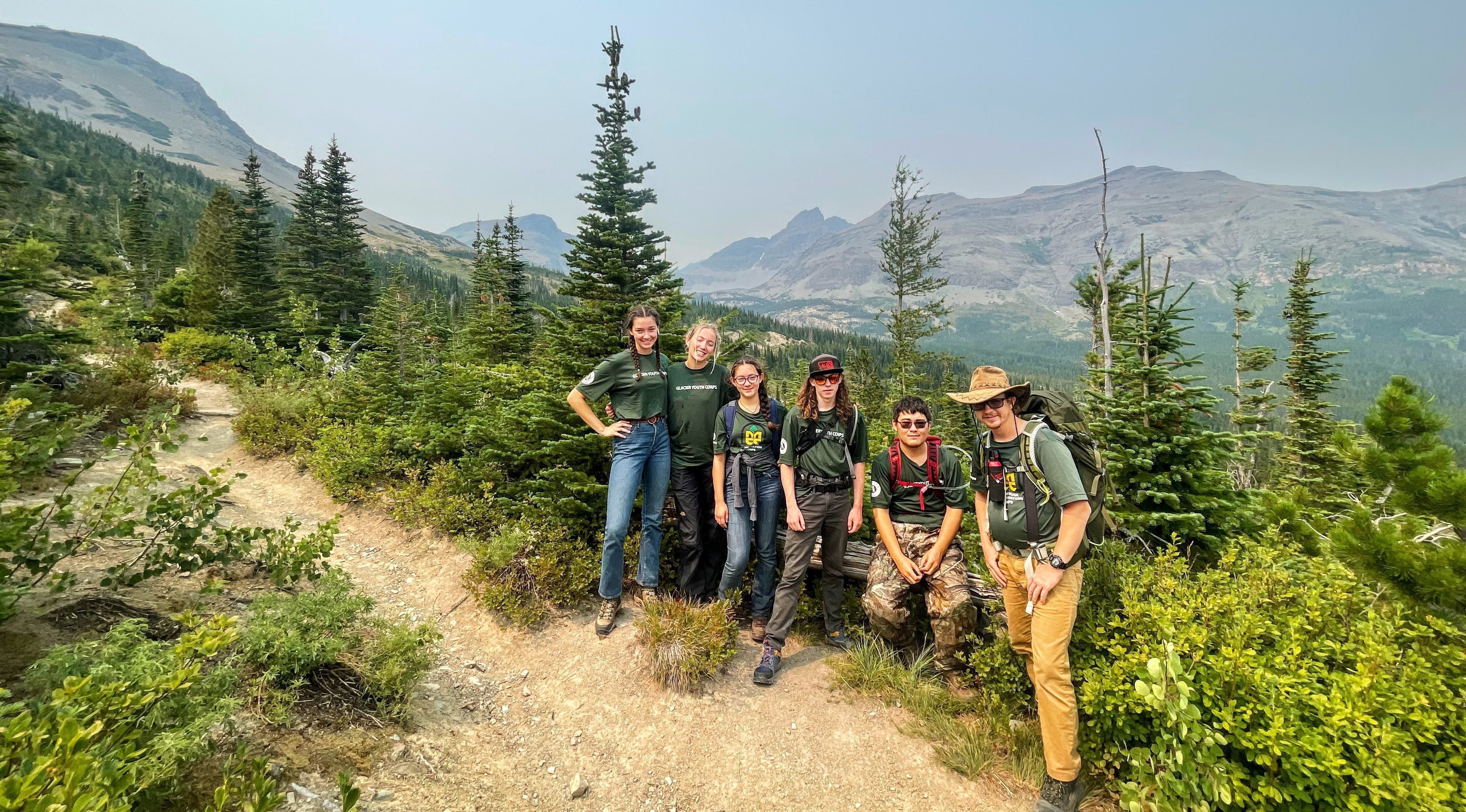 [Image Description: Six MCC members standing together on a trail, overlooking a mountainous landscape, surrounded by green shrubs and pine trees.]