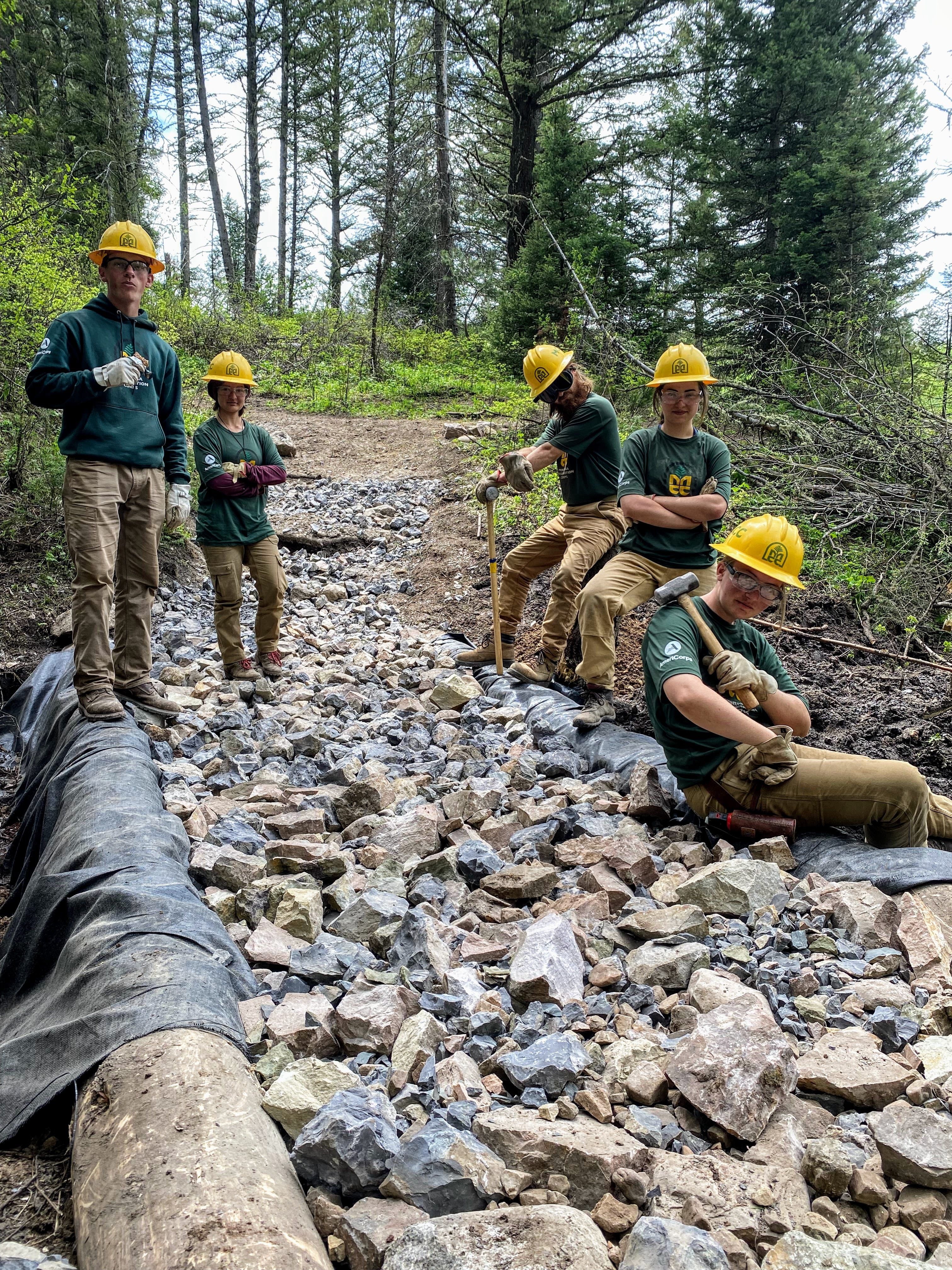 A crew stands slightly uphill of the camera, clustered around the edges of a rocky trail. There are tarp covered logs bordering the rocky trail on both sides.
