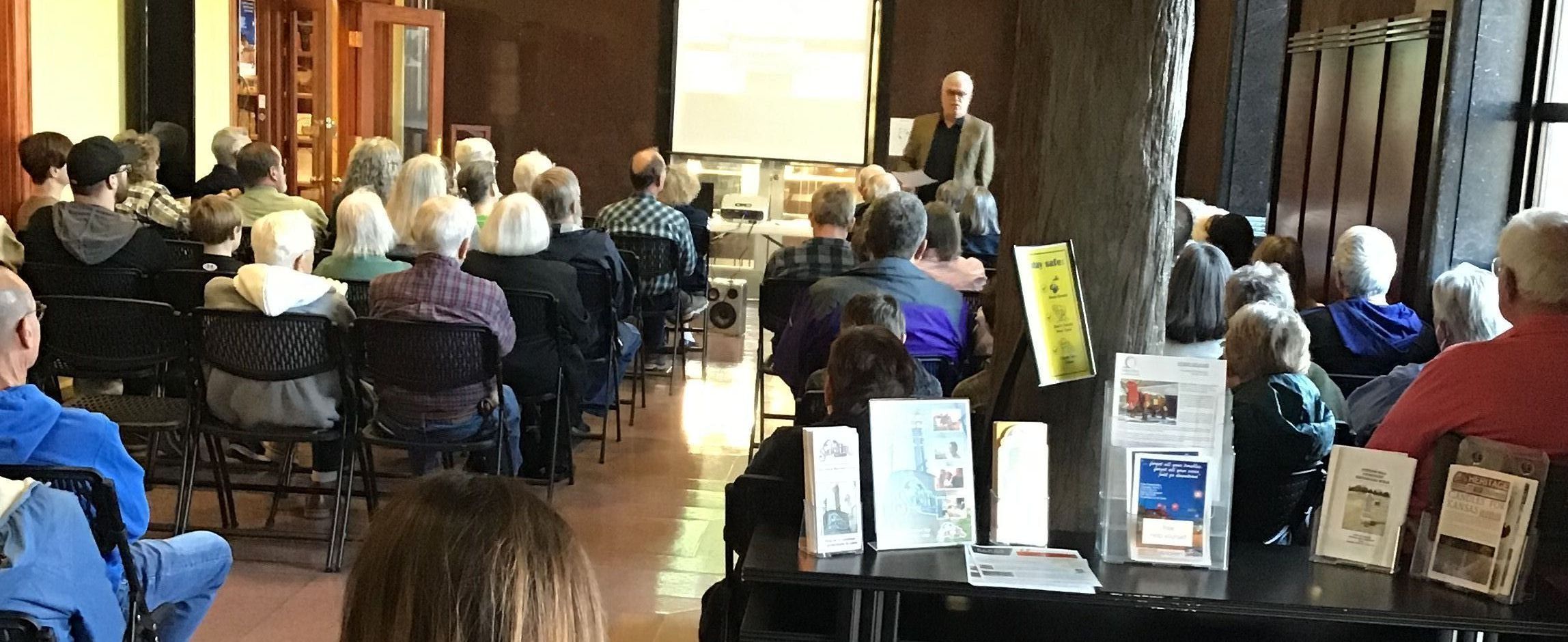 Group of a lot of people sitting in the Smoky Hill Museum lobby facing a male speaker giving a presentation.