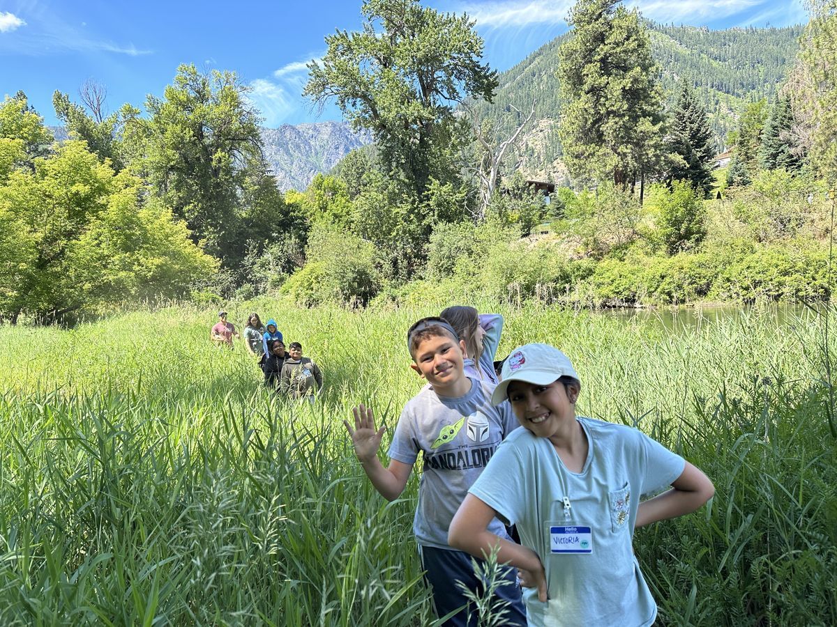 A group of students in a meadow with tall grass and mountains in the background.