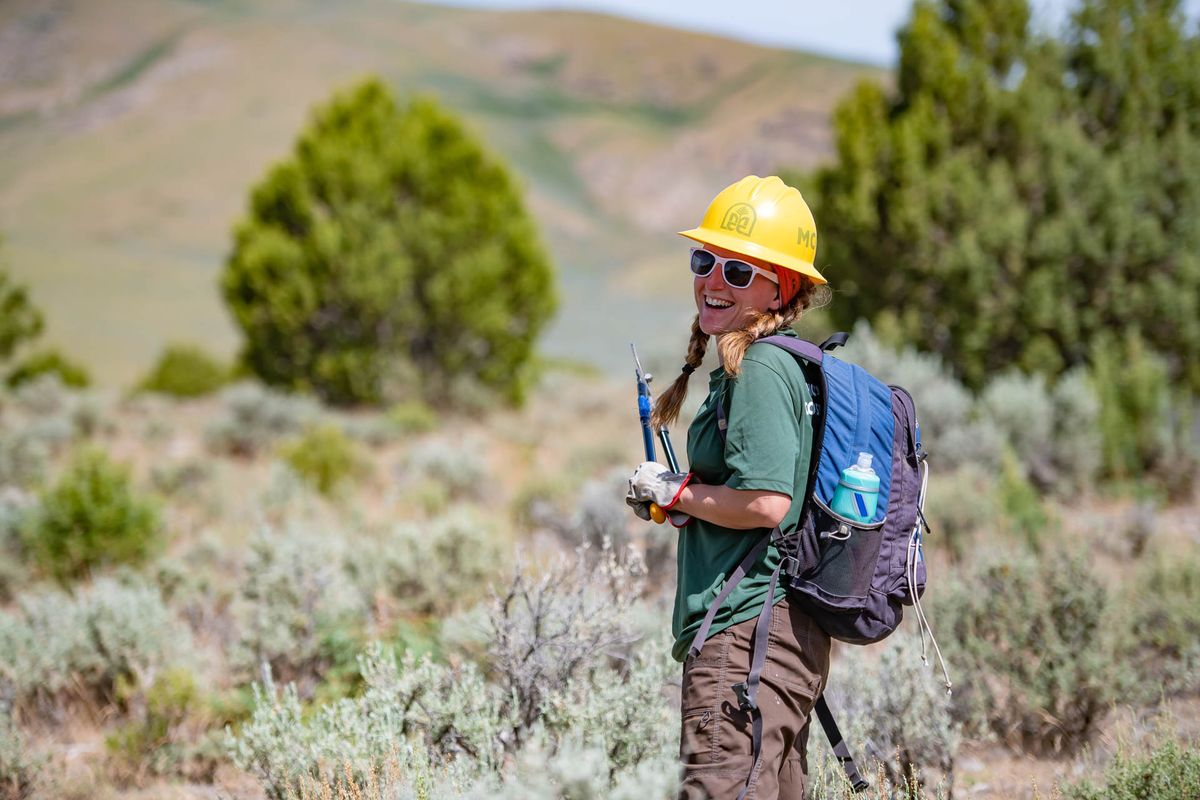 [Image Description: An MCC member standing in a sagebrush field, smiling holding a clipboard, wearing a hard hat and back pack.]