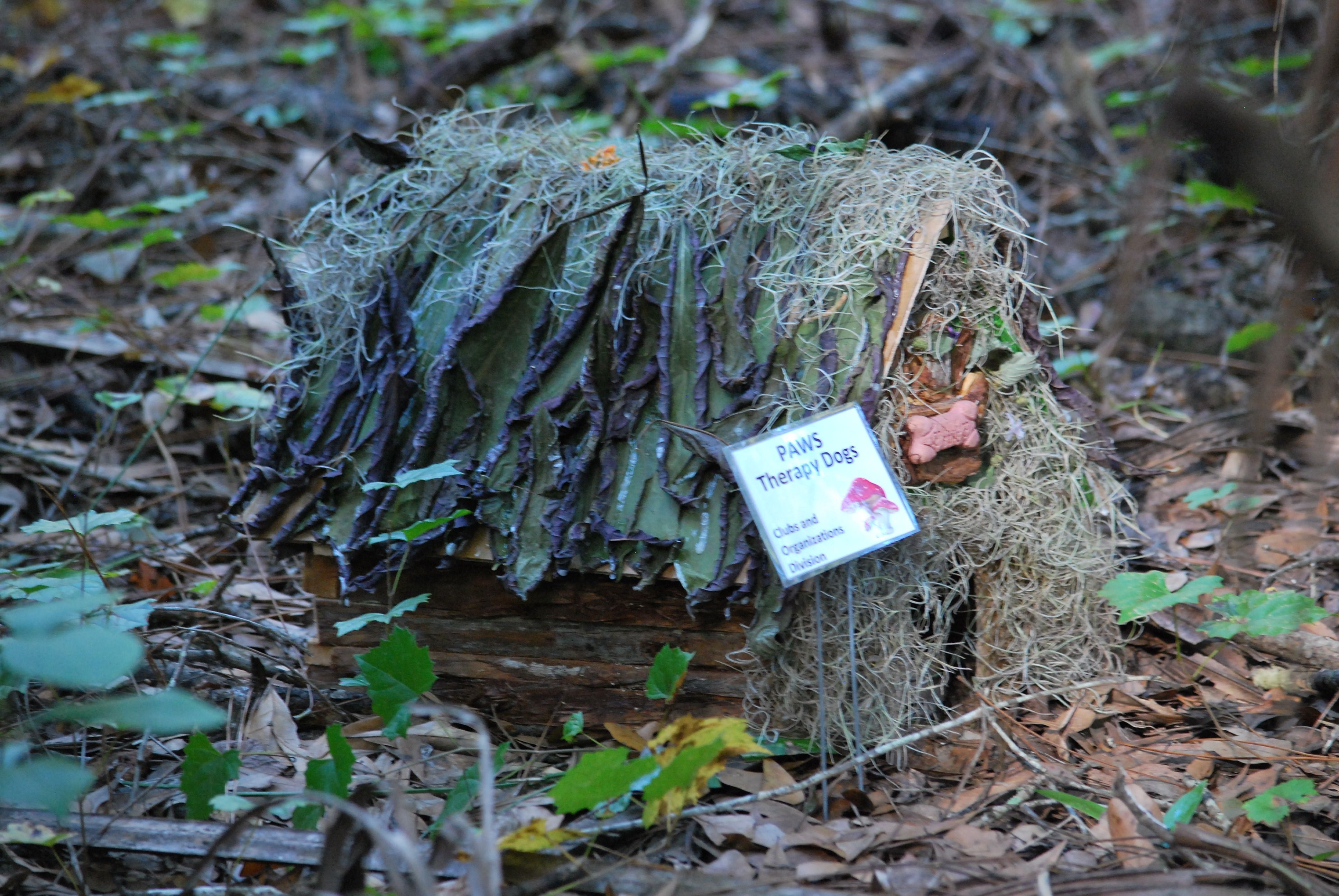 Nestled in leaves and grape vine is a small wooden house with slanted roof decorated with Spanish moss. It has a small dog bone over the door representing the PAWS therapy dog organization. 