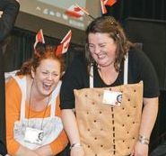 Two girls stand side-by-side laughing. They both are in costume with Canadian flags in their hair. One girl is dressed like a graham cracker and the other is dressed like a marshmallow.