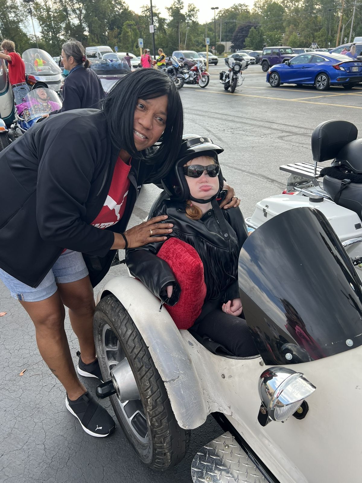 A young woman sits in a motorcycle sidecar wearing a helmet and looking toward the camera.  A woman older than her stands behind her, bending over with her hands on the woman's shoulders and smiling.