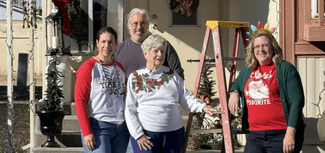Three volunteers-one man and two women standing with homeowner after decorating the outside of her home with holiday lights