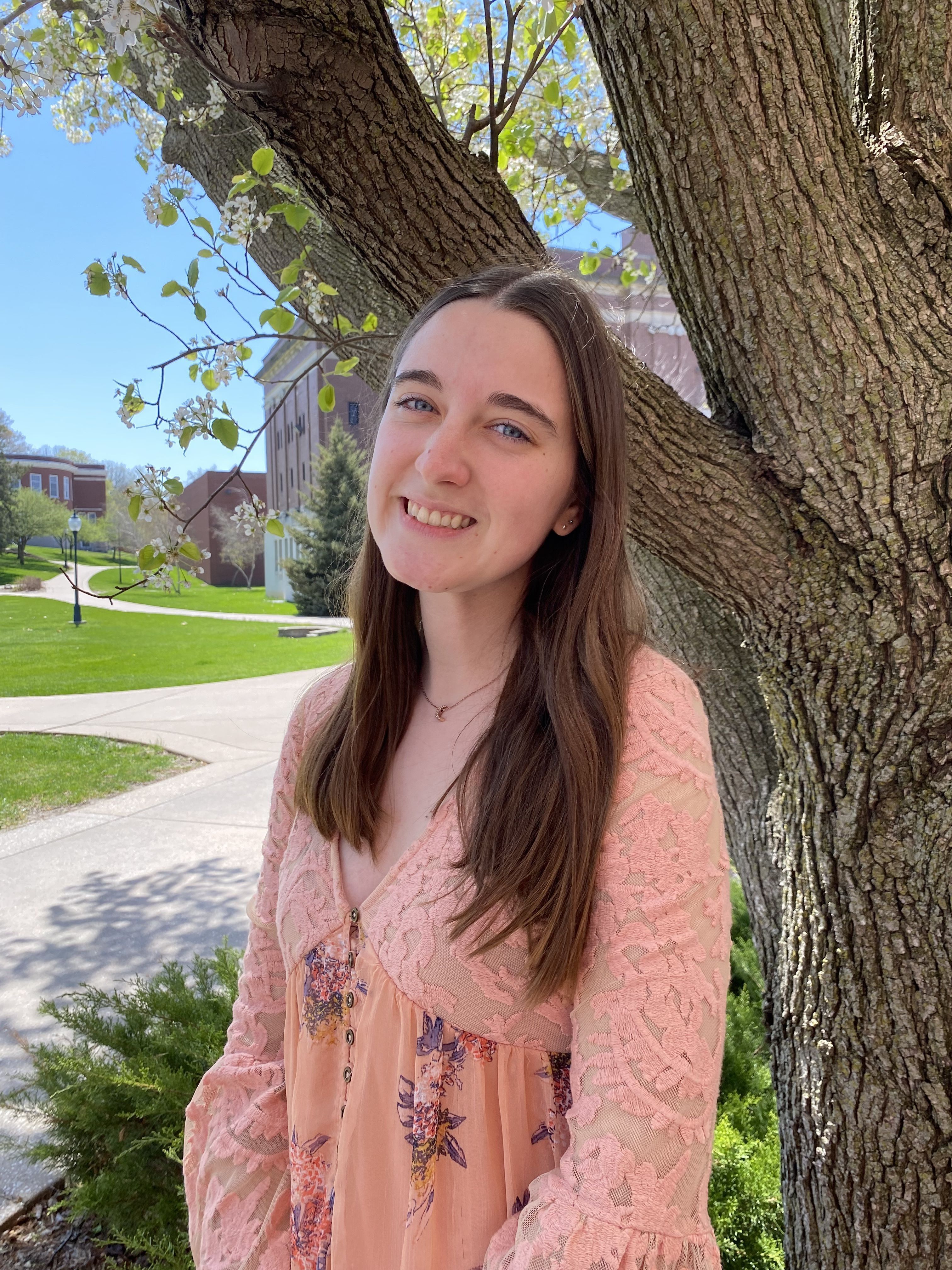 Bailey Hill, a young woman with long brown hair, smiles while standing outdoors next to a tree. She is wearing a peach and pink floral dress with lace sleeves. The background features a college campus with green lawns, brick buildings, and a bright blue s