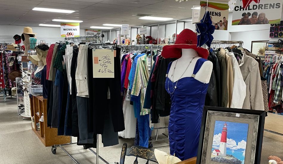 Photo of thrift store's interior, showing mannequin in a blue cocktail dress and red wide brimmed hat. Tidy racks of clothes, hats, sunglasses, and home decor stretch into the distance.