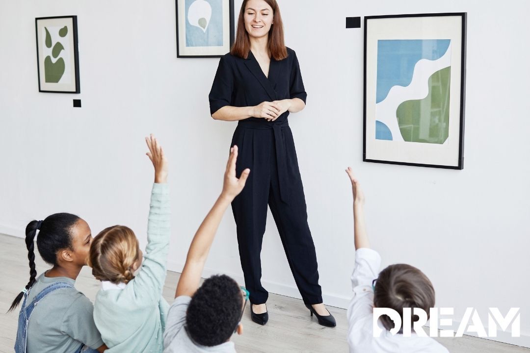 Woman standing at art gallery in front of a few sitting kids with their hands raised