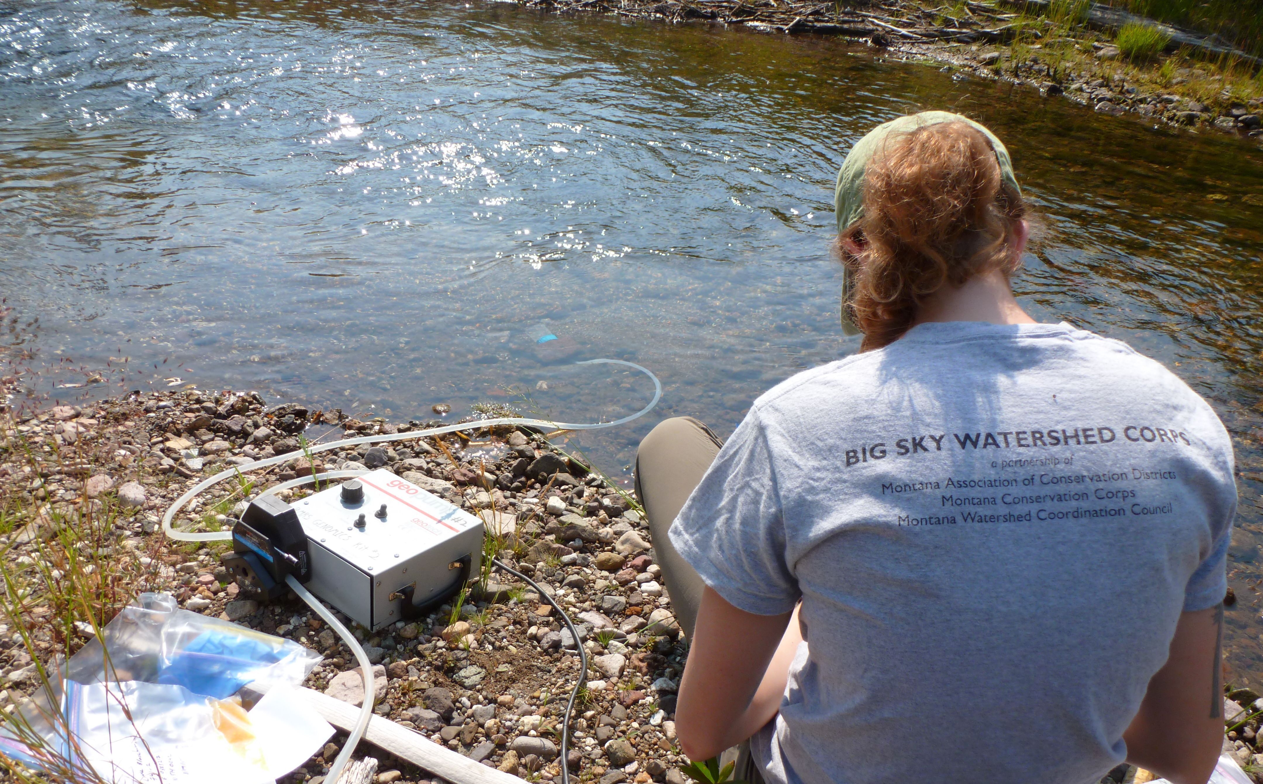 [Image Description: A Big Sky Watershed Corps member sits on a streambank, collecting samples of the water.]