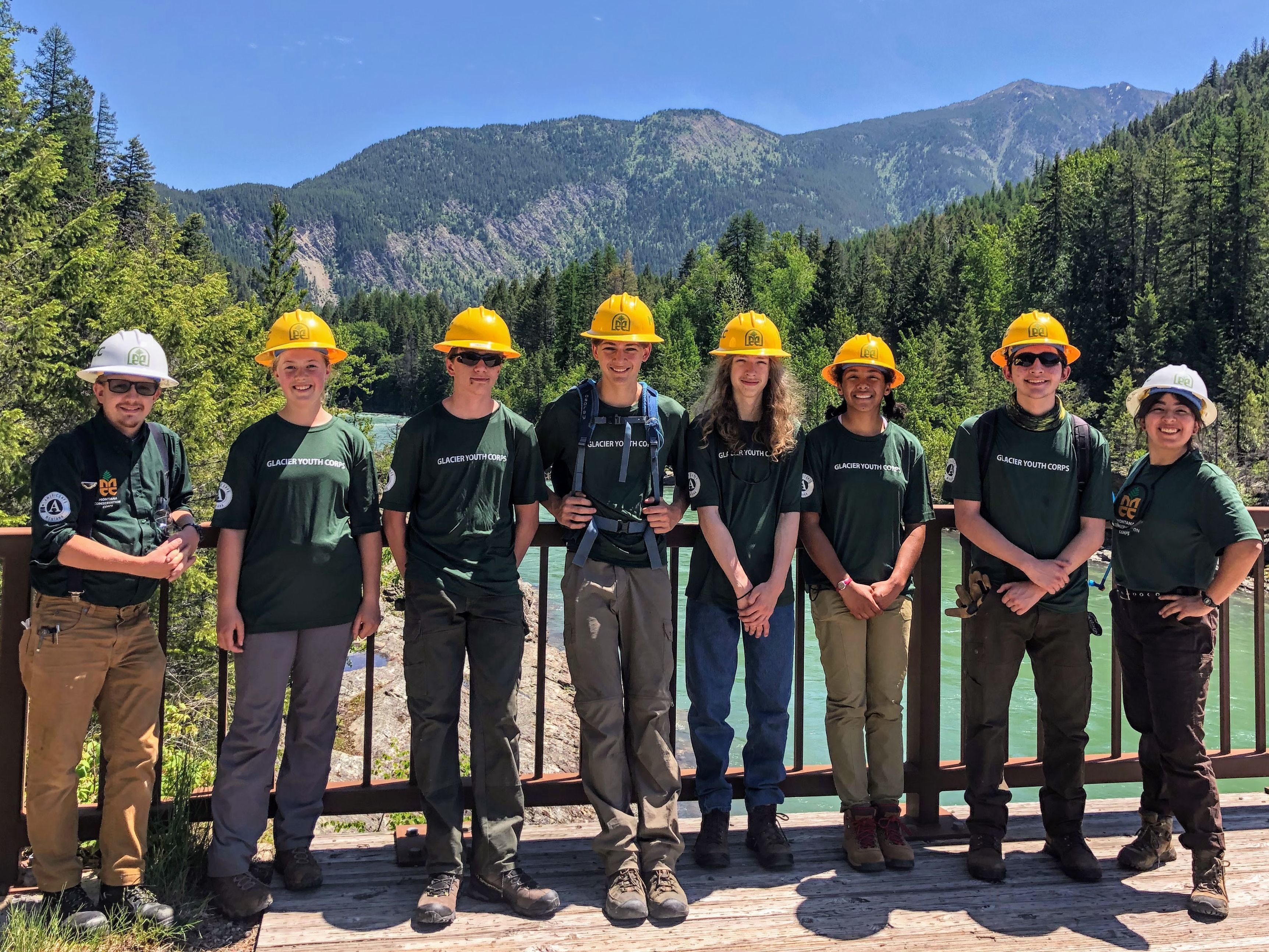 [Image description: A youth crew stands and two leaders stand on a bridge with mountains in the background. Everyone is wearing MCC uniforms]