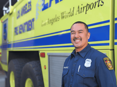 Firefighter/Engineer Ronald Tomacruz smiling and standing in his blue uniform front of the specialized, large, green Fire Station 80 rig.