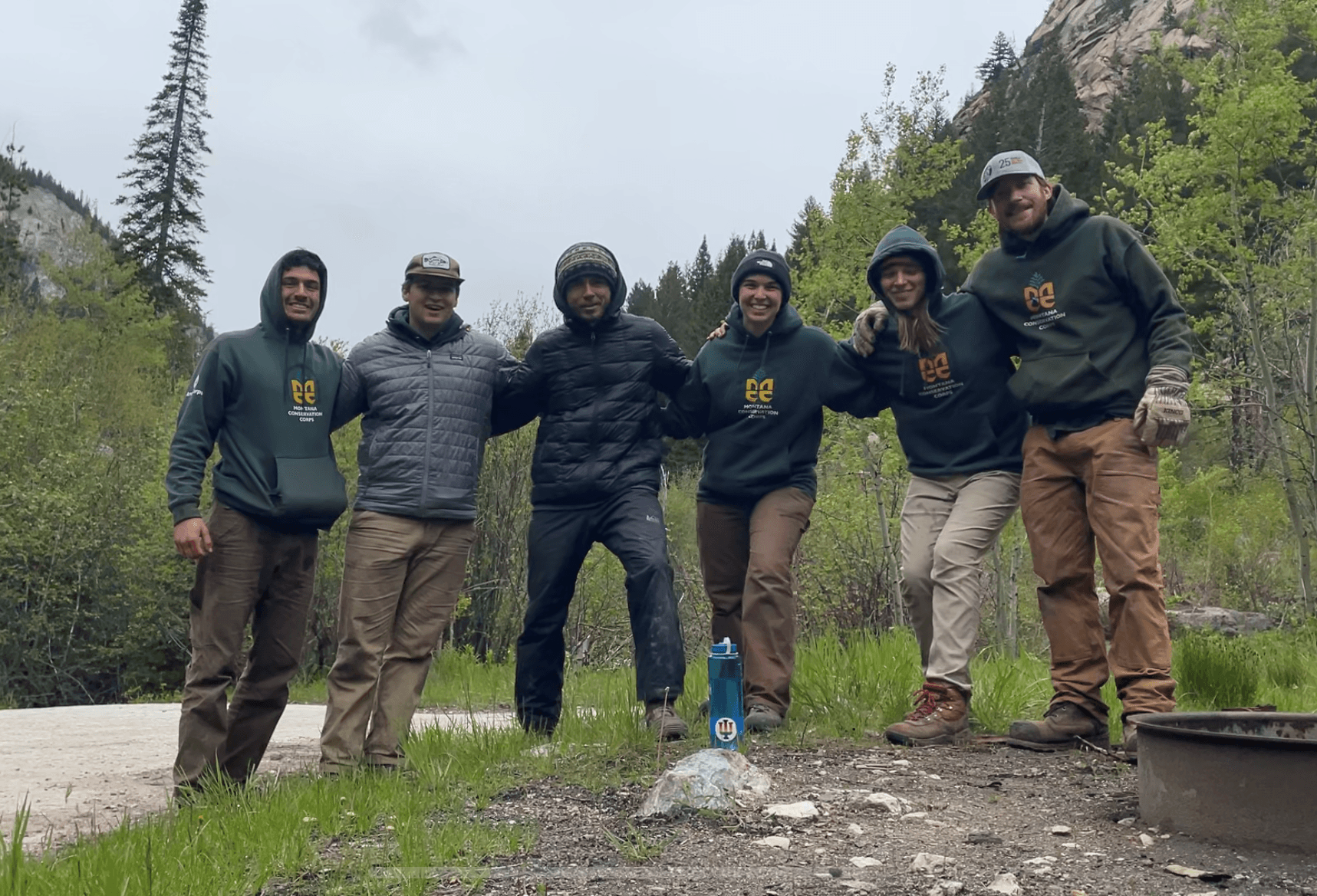 A crew poses together behind a fire ring. They are all bundled up in the cold and their is overcast sky and a hillside with trees and undergrowth behind them.