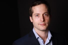 Stuart Green. A man who is in his 30's and he is wearing a light blue shirt with a blazer. He is posing for the picture. There is a dark background behind of him while he has a spotlight on him.