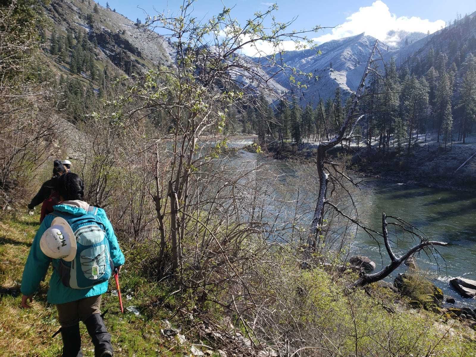 A crew walks along side the Salmon River. There are snow capped mountains in the distance.