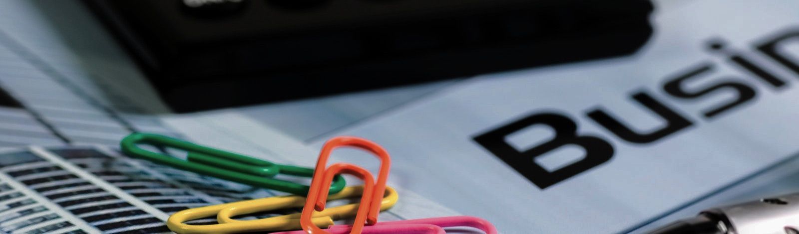 Closeup photo of miscellaneous paperwork and desk supplies, including colorful paperclips, charts, the bottom edge of a calculator, and the word "Business." The colorful paperclips are in the left half of the photo in the foreground and are orange, yellow