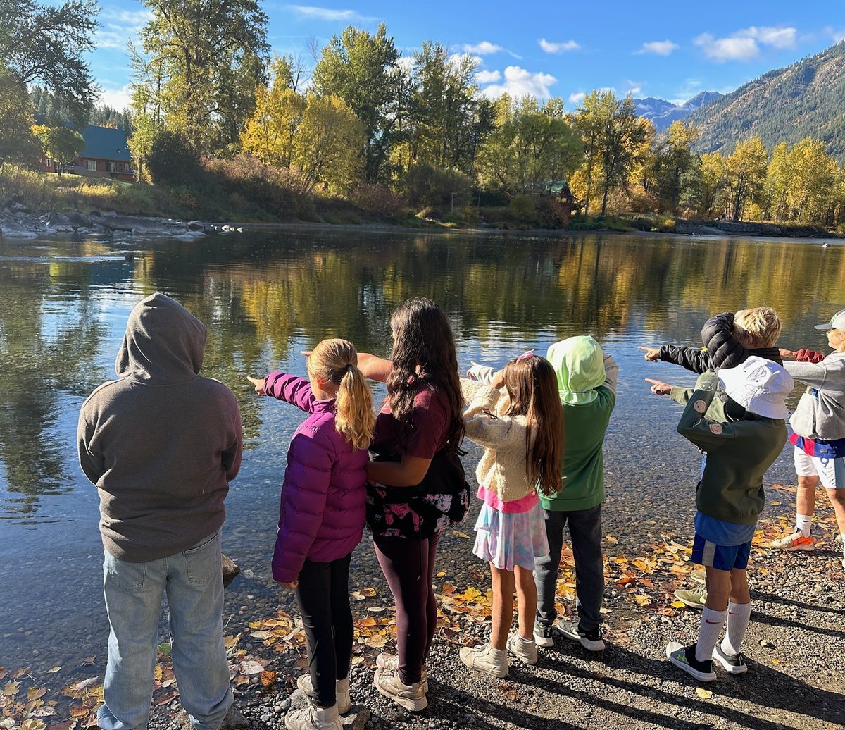 A group of students on the bank of the Wenatchee River pointing at a fish, surrounded by the fall foliage.