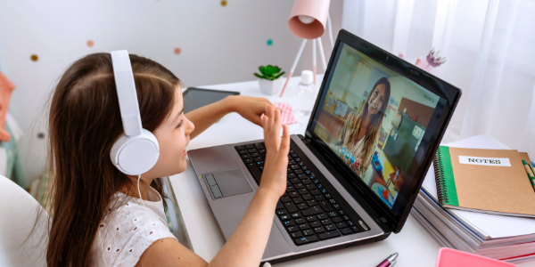 A young girl with long brown hair wears white headphohnes and gestures at her laptop screen as her teacher smiles. She is learning via zoom!