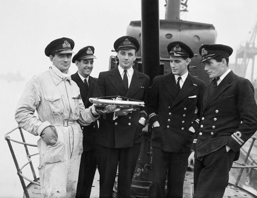 A B&W photo of five young male naval officers are depicted here on the deck of their submarine in military uniforms. One on the far left wears a light color, the others wear dark. The man in light color holds a model boat toward the center of the group.