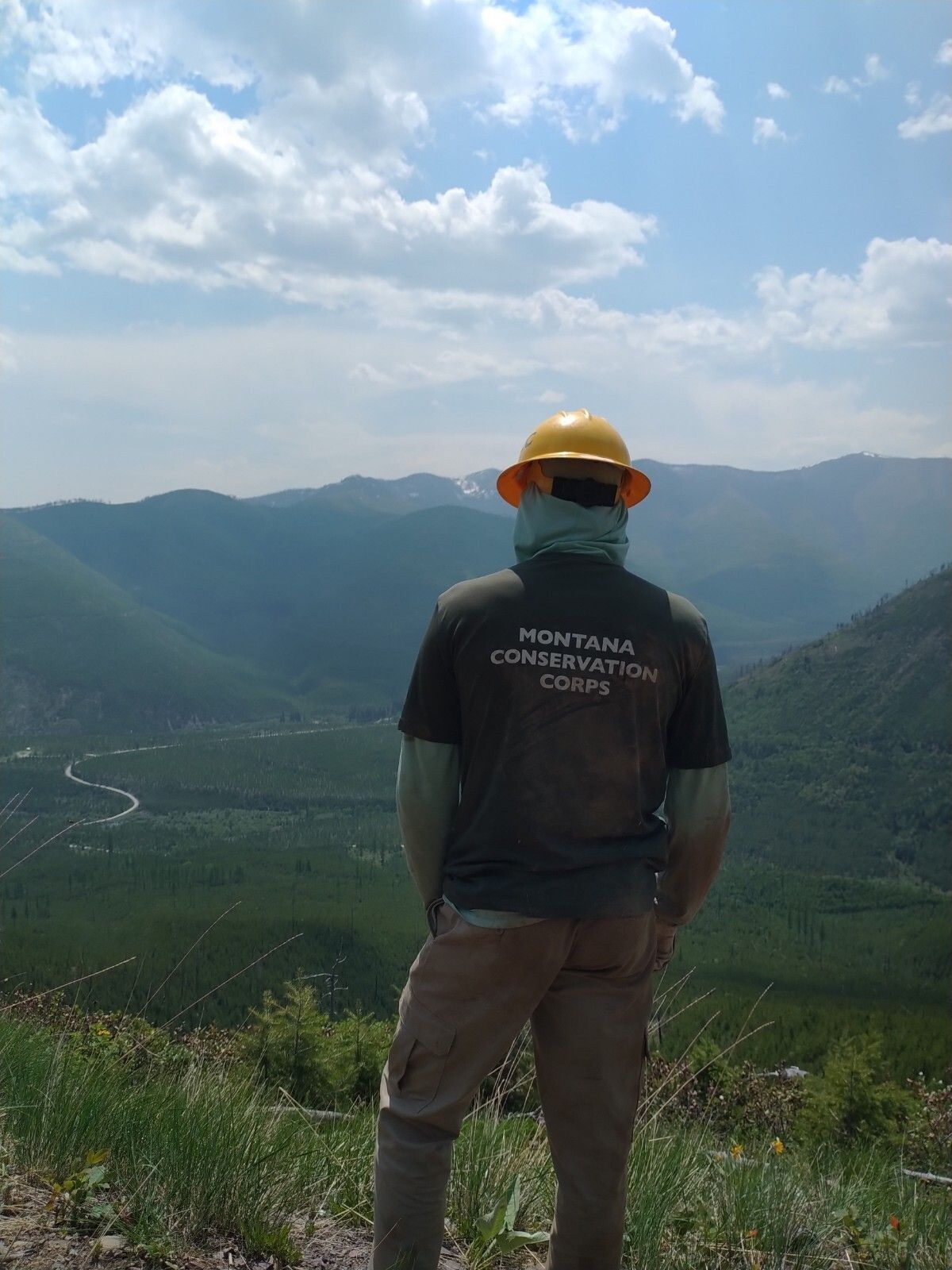 A crew member wearing an MCC shirt and helmet faces away from the camera, looking down into a valley.