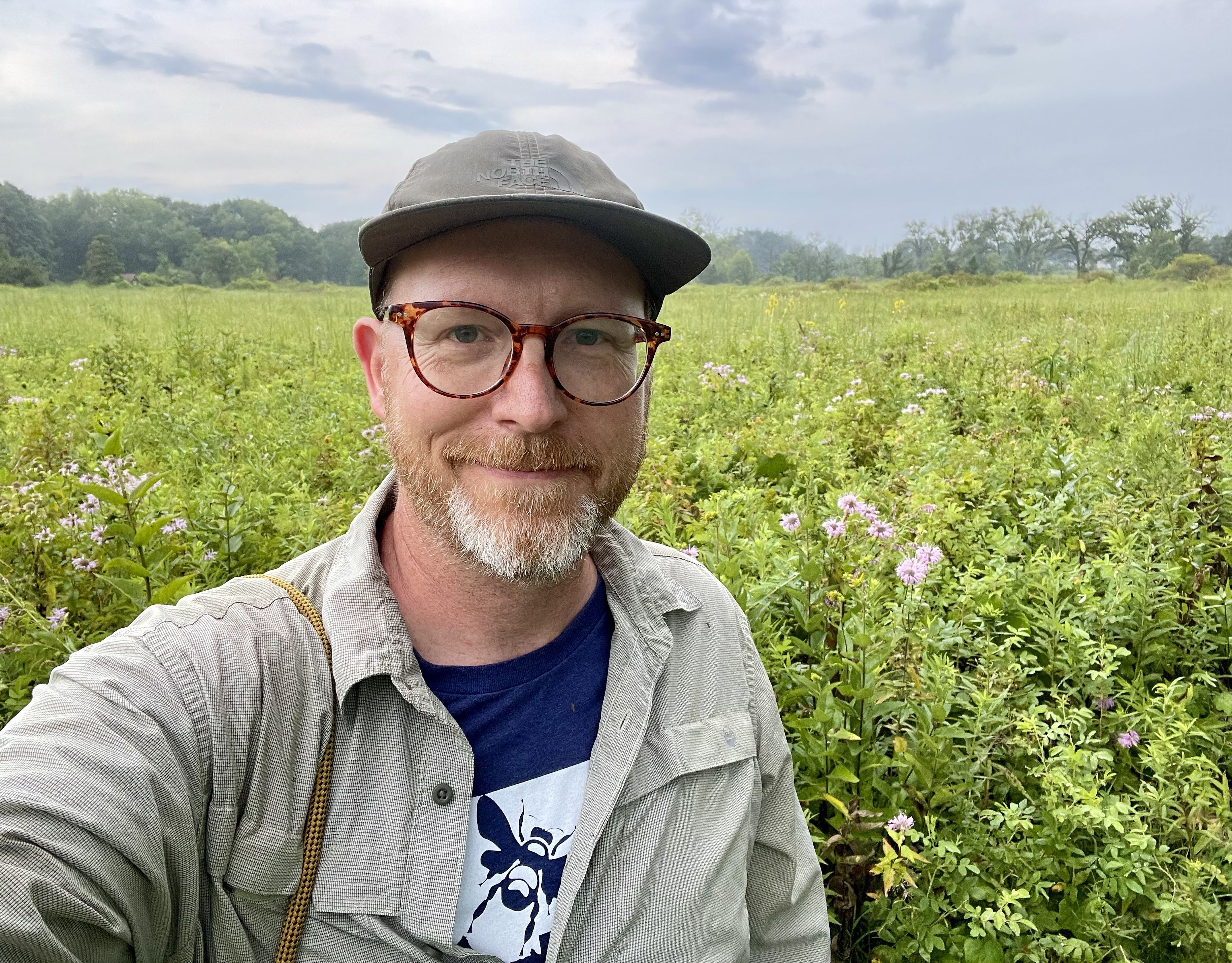 man in a hat and bumblebee shirt stands in a field of flowers