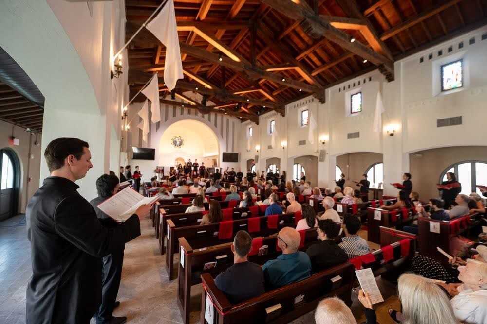 Singers line up around the perimeter of a church sanctuary, surrounding a full audience from all sides.