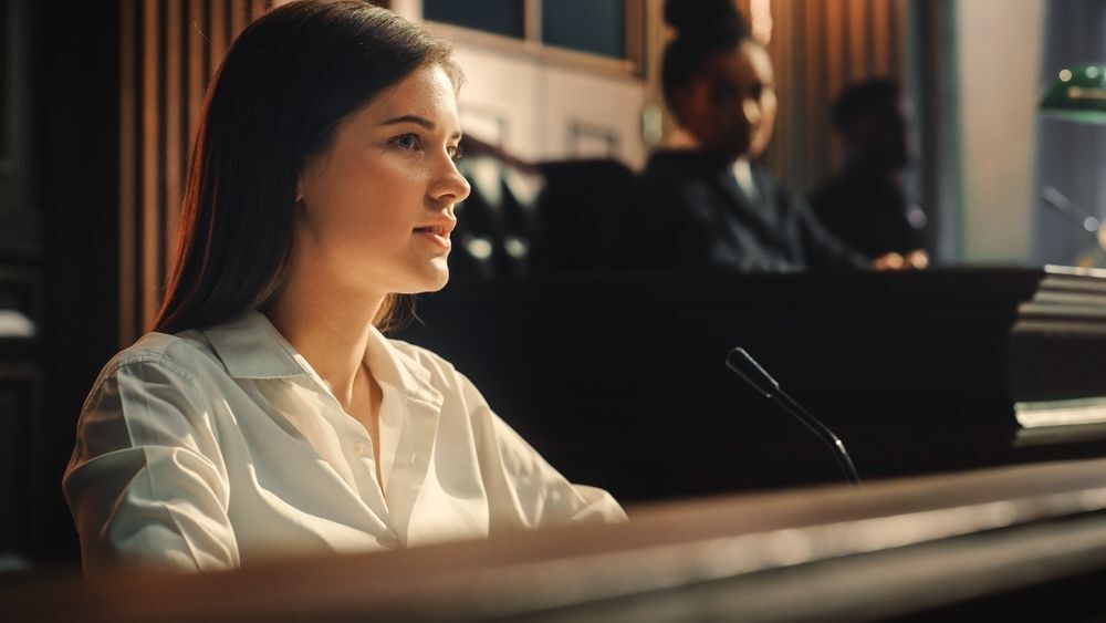 Image of a woman in professional attire taking the stand at a hearing in a court room. Behind her is the judge listening attentively. 