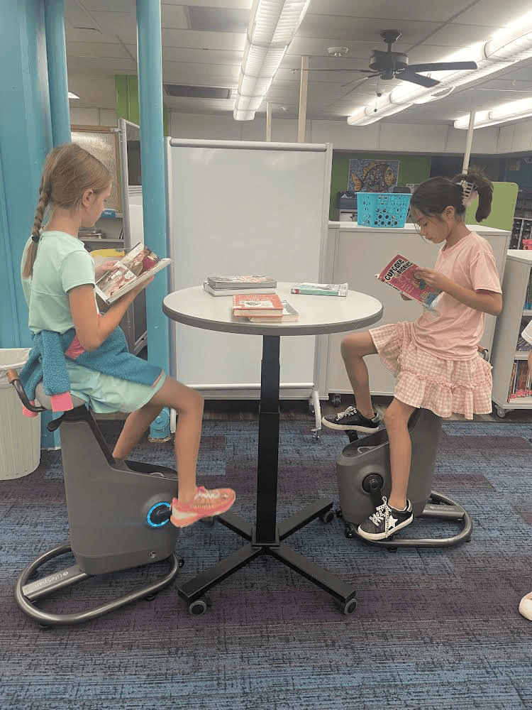  Two students sit on stationary bikes in a library, reading books while pedaling. The bikes are placed around a small table with more books, creating a calm and focused learning environment.